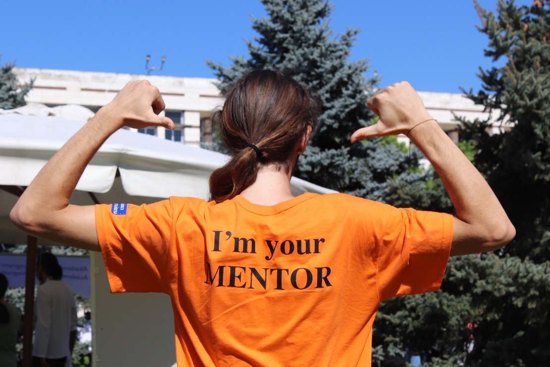 Turuncu "I'm your MENTOR" tişörtüyle sırtını gösteren bir mentor öğrenci
